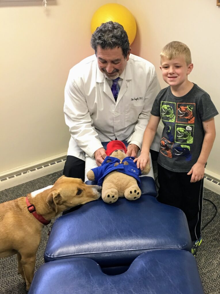 Dr. Steve Segal adjusts a teddy bear on a chiropractic table while smiling with a young boy and his dog, Charlie, in the Libertyville clinic.